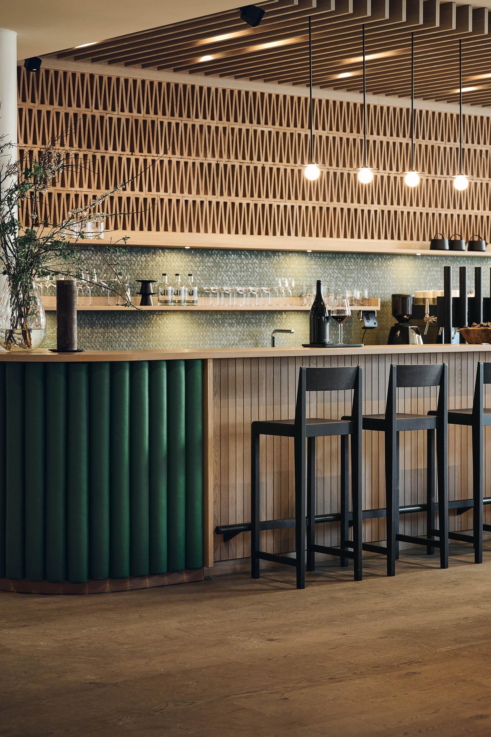 An elegant bar with green design and minimalist wooden cladding. Bright pendant lights hang over the high chairs that invite sitting.