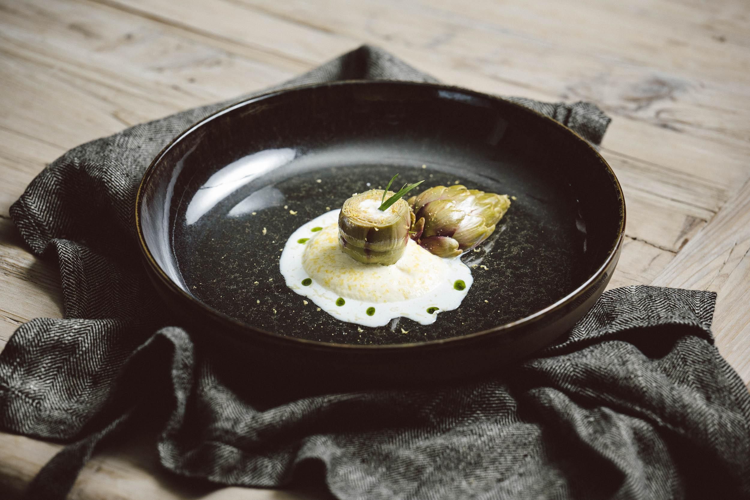 An elegant dish on a black plate with a creamy sauce. The presentation features artistically arranged ingredients and fresh herbs.