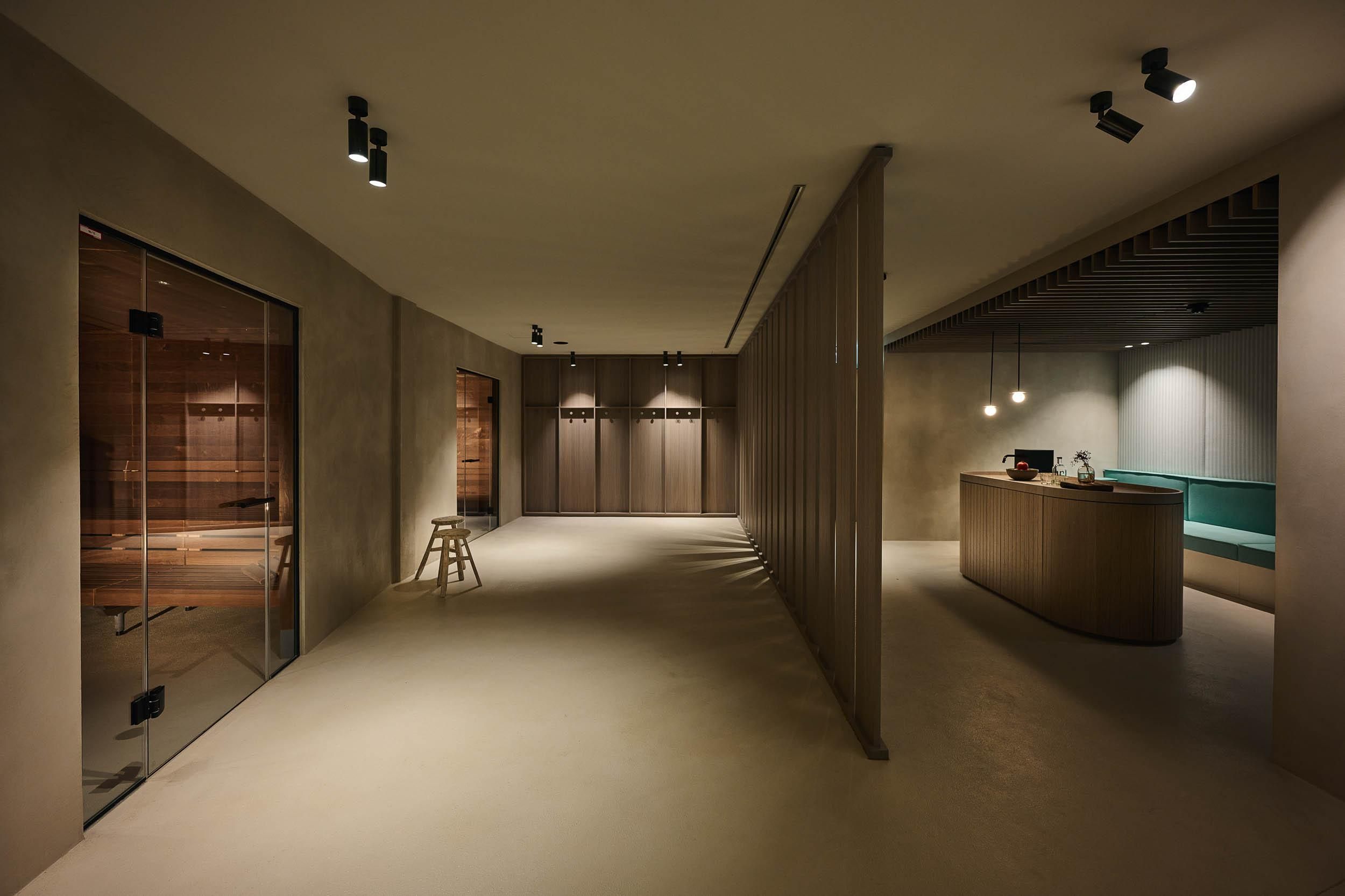 A modern, minimalist reception area with soft colors and subdued lighting. On the left side, there are glass doors that lead to a sauna.