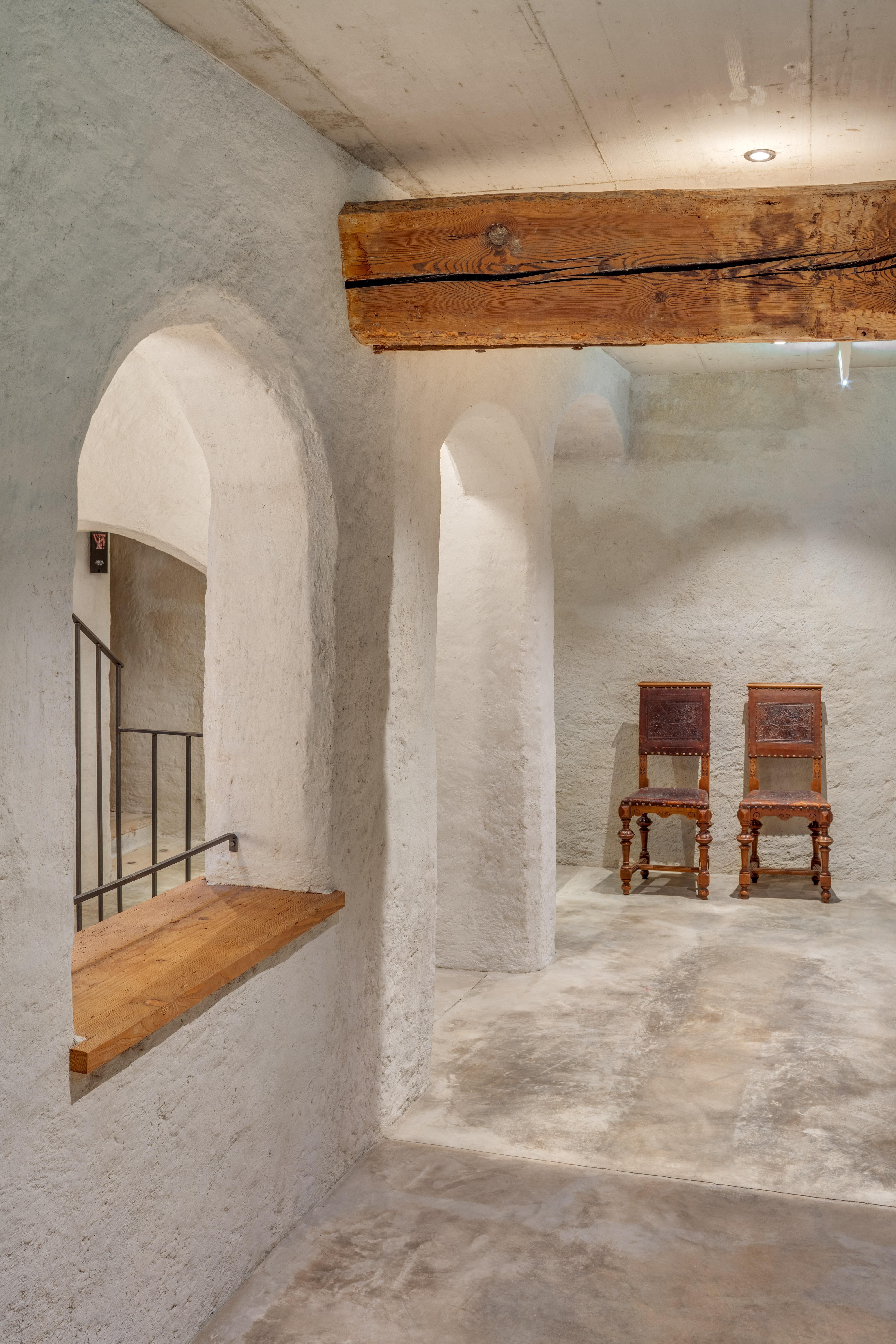 A bright room with a continuous wall of white plaster. Two rustic wooden chairs face each other.