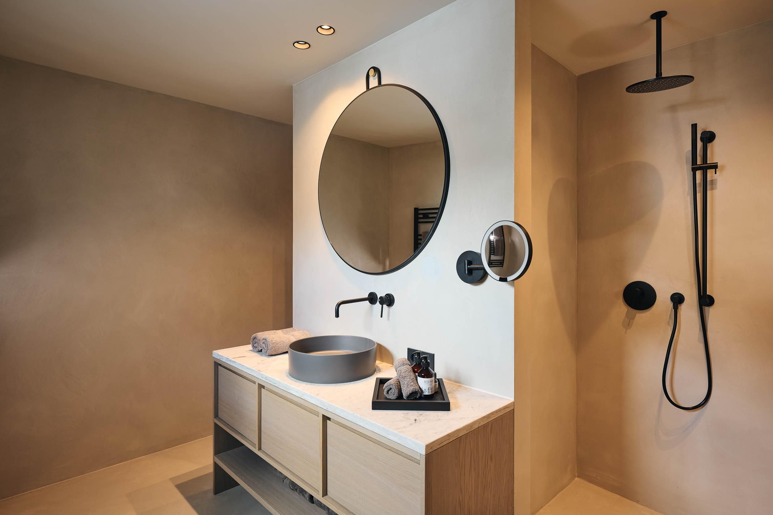 A modern bathroom with a round mirror and an elegant sink unit. The walls are in neutral colors, and there is a shower with a black showerhead.
