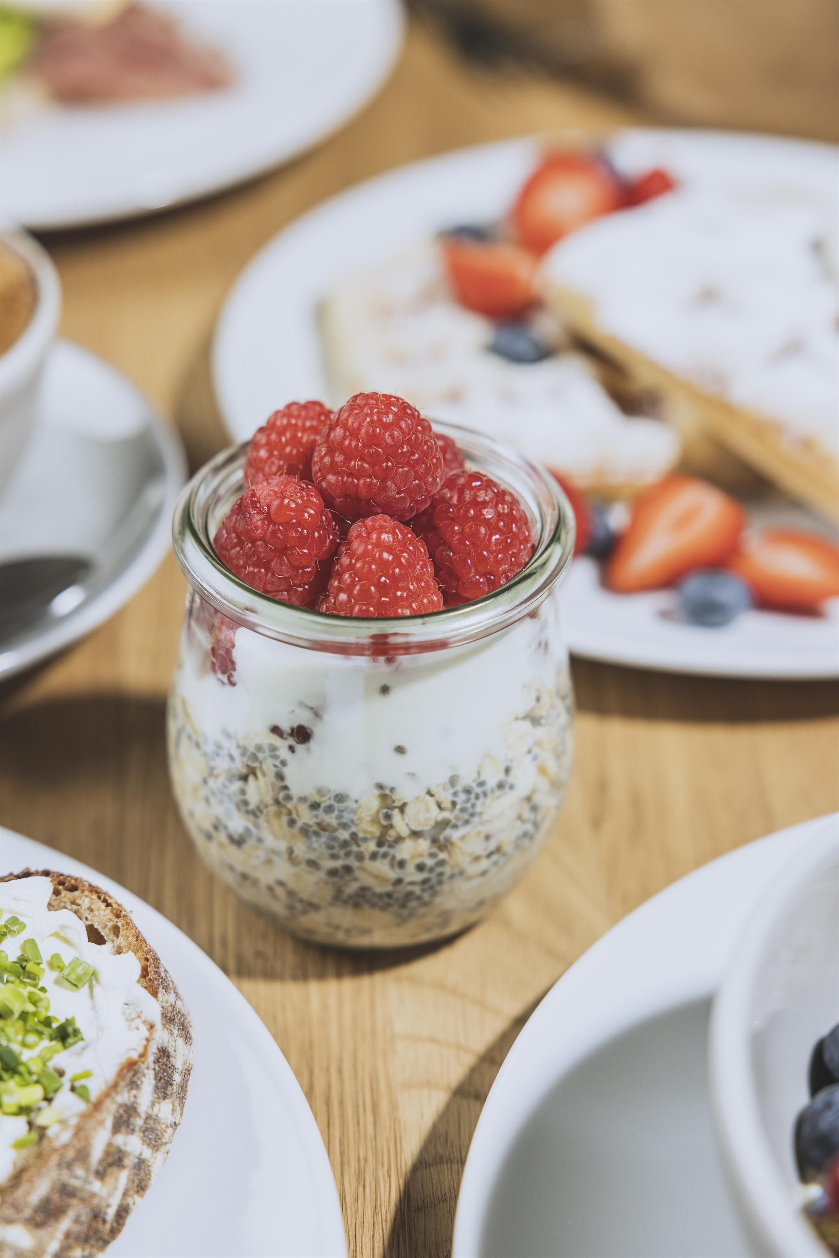 A glass filled with yogurt, oats, and fresh raspberries is placed on a wooden table. In the background, there are additional plates with various fruits and snacks visible.