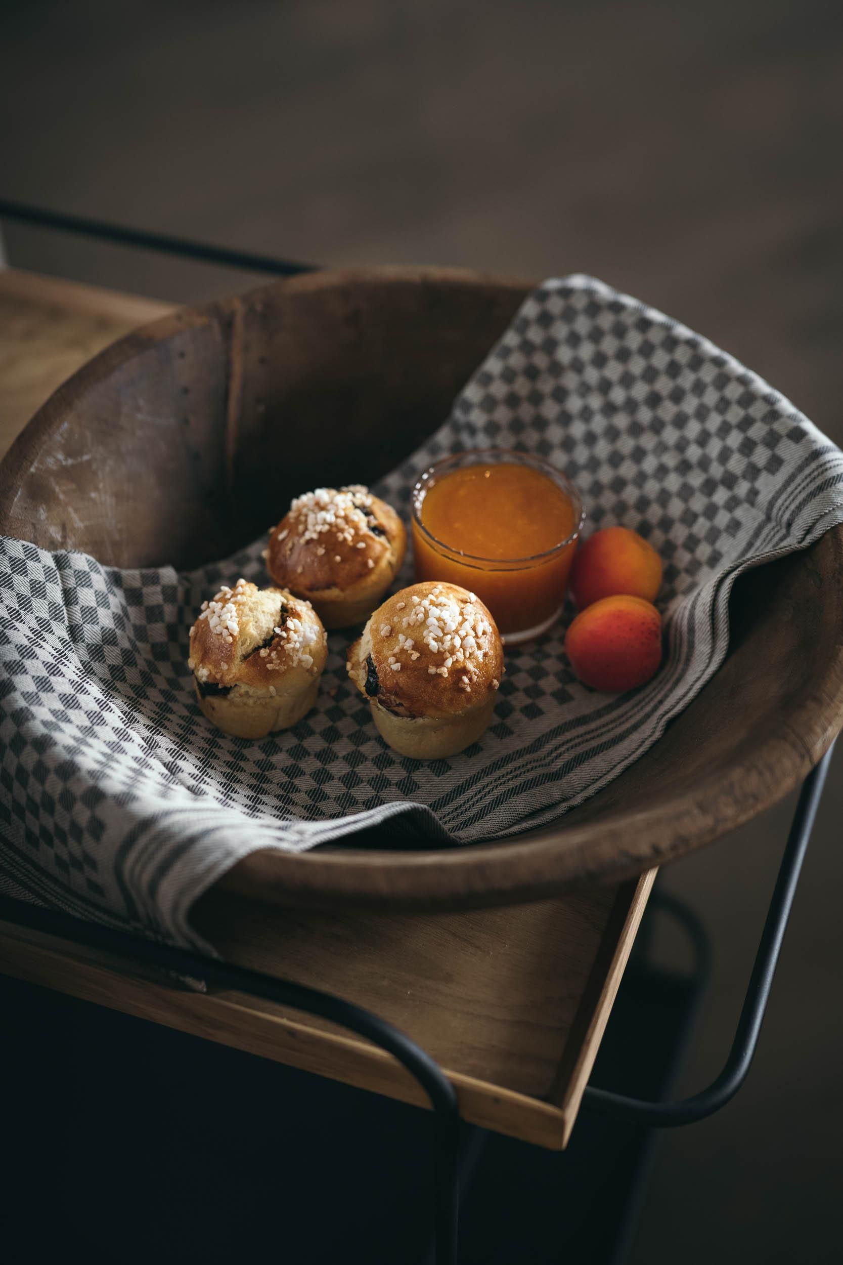 A wooden bowl with three pastries that are decorated with sprinkles. There is also a glass with an orange liquid and some fruits.